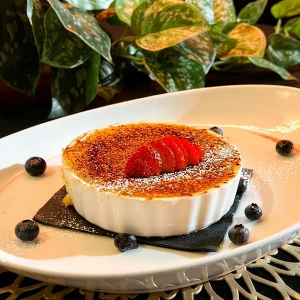 Crème brûlée in a white ramekin, topped with sliced strawberries and powdered sugar, garnished with blueberries.