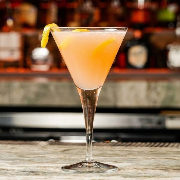 A pink cocktail in a martini glass with a lemon twist sits on a marble bar.