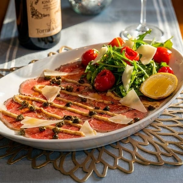 Carpaccio with capers, parmesan, arugula salad, cherry tomatoes, and lemon on a white platter.