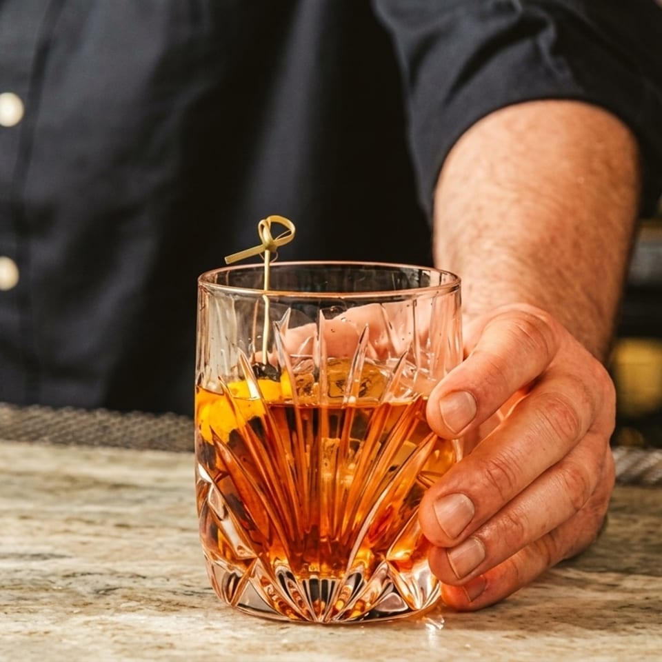 Bartender's hand holding a crystal glass with an amber cocktail and orange peel garnish.